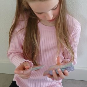 Een meisje met een zachtroze trui houdt een stapel affirmatiekaarten voor kinderen vast in haar ene hand. In de andere hand heeft ze een los affirmatiekaartje. De affirmatiekaartjes hebben een pastelkleurig ontwerp.