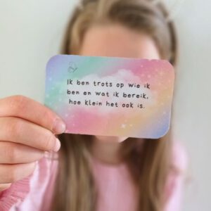 Een meisje houdt een affirmatiekaart voor kinderen vast met de tekst: "Ik ben trots op wie ik ben en wat ik bereik, hoe klein het ook is." De achtergrond van de affirmatiekaart voor kinderen heft een pastel regenboogkleurig verloop.