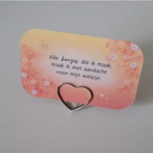Een affirmatiekaart met een Nederlandse tekst, gericht op persoonlijke ontwikkeling en zelfreflectie. Het affirmatiekaartje is geplaatst in een hartvormig houdertje en heeft de kleuren oranje en geel met een bloesemontwerp.