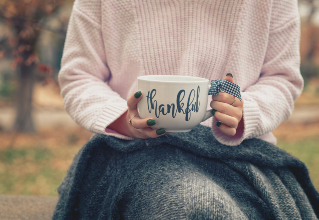 Een vrouw met groene nagellak houdt een witte mok vast met woord `thankful` erop geschreven. De mok wordt vastgehouden met beide handen, en er is een zwart/wit geruite strik om oor van het kopje gebonden. de achtergrond is wazig en de vrouw zit onder een grijze deken.